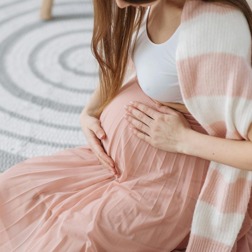 Pregnant woman sitting indoors, tenderly holding her baby bump in soft, cozy attire.